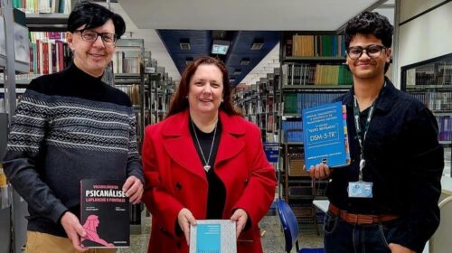 Na biblioteca da FCM, três pessoas estão em pé atrás de uma mesa redonda onde estão expostos vários livros de Psicologia Médica e áreas relacionadas. À esquerda, um homem de suéter cinza e óculos segura o livro Vocabulário da Psicanálise. Ao centro, uma mulher de casaco vermelho segura um exemplar de Psicologia Médica. À direita, um jovem de crachá e camisa escura segura o Manual Diagnóstico e Estatístico de Transtornos Mentais – DSM-5-TR. Ao fundo, há estantes repletas de livros.