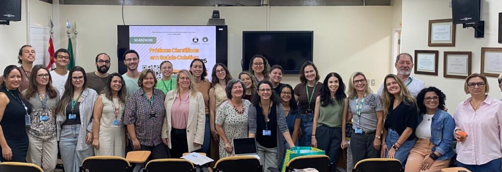A imagem é um registro de grupo em uma sala de aula, mostrando uma grande quantidade de pessoas, a maioria mulheres, em pé e sorrindo. O grupo parece ser composto por estudantes e professores. No fundo da sala, uma tela de projeção exibe o tema do evento, "Práticas Científicas em Saúde Coletiva 2025", e o logo da UNICAMP. No lado direito, em uma parede, há vários quadros com diplomas ou certificados pendurados, e uma bandeira do estado de São Paulo está visível à esquerda.