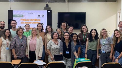 A imagem é um registro de grupo em uma sala de aula, mostrando uma grande quantidade de pessoas, a maioria mulheres, em pé e sorrindo. O grupo parece ser composto por estudantes e professores. No fundo da sala, uma tela de projeção exibe o tema do evento, "Práticas Científicas em Saúde Coletiva 2025", e o logo da UNICAMP. No lado direito, em uma parede, há vários quadros com diplomas ou certificados pendurados, e uma bandeira do estado de São Paulo está visível à esquerda.