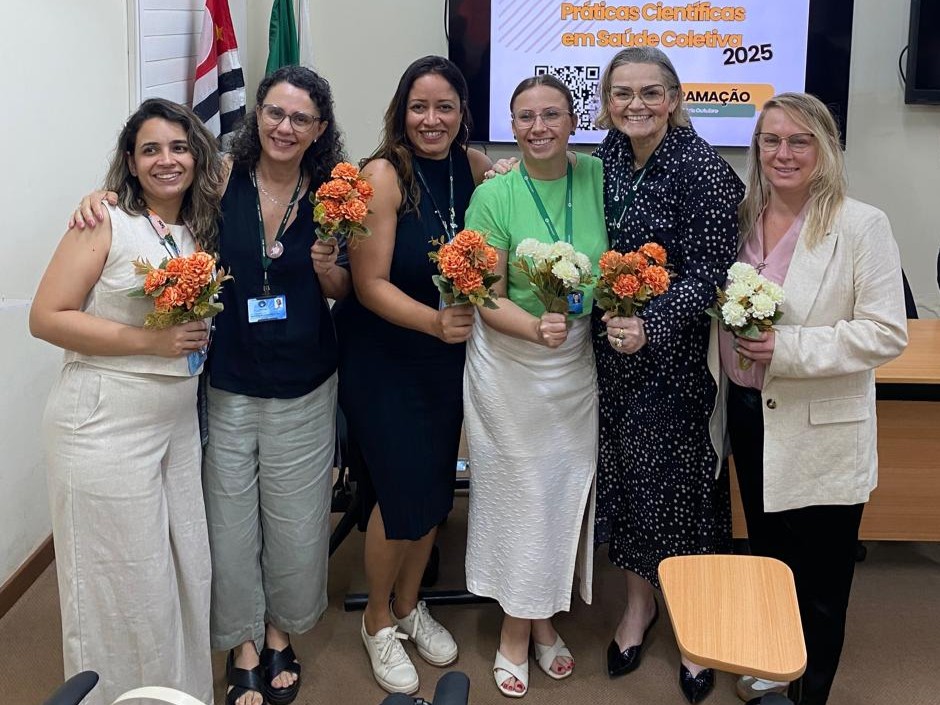 A fotografia registra um grupo de seis mulheres sorrindo, posando lado a lado em um ambiente interno que parece ser uma sala de reuniões ou aula. Elas estão abraçadas ou com os braços nos ombros umas das outras e todas seguram pequenos buquês de flores (laranja e brancas). Ao fundo, uma tela de televisão exibe parcialmente o texto "Práticas Científicas em Saúde Coletiva 2025".