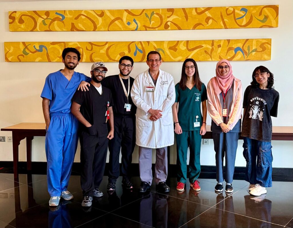 Seis estudantes de Medicina e um médico posam sorridentes em um corredor do Hospital Hamad, no Catar. Eles usam jalecos e roupas hospitalares, e atrás deles há três painéis amarelos com desenhos abstratos.