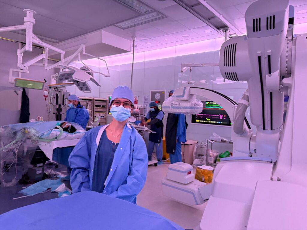Estudante de Medicina com máscara e roupa cirúrgica azul acompanha procedimento no centro cirúrgico do Hospital Hamad, no Catar. Ao fundo, equipe médica trabalha ao redor do paciente e monitores exibem dados vitais.
