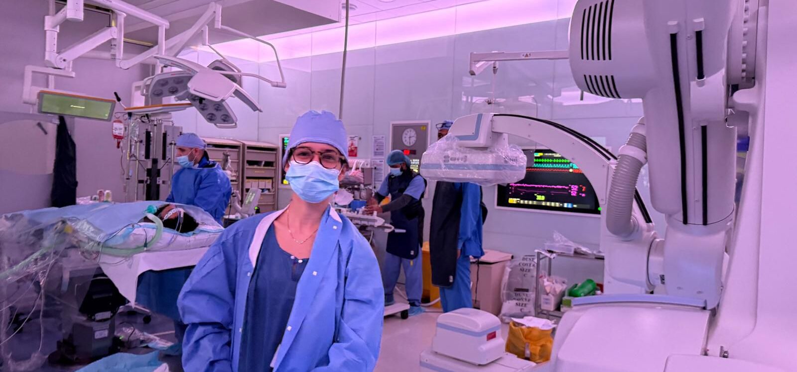 Estudante de Medicina com máscara e roupa cirúrgica azul acompanha procedimento no centro cirúrgico do Hospital Hamad, no Catar. Ao fundo, equipe médica trabalha ao redor do paciente e monitores exibem dados vitais.
