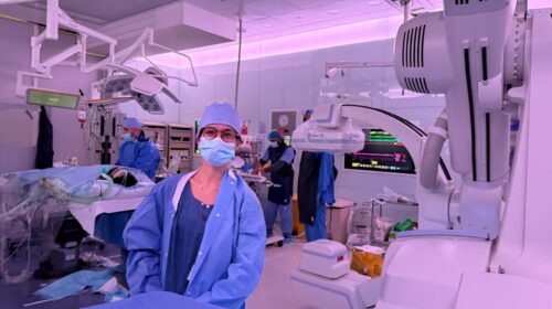 Estudante de Medicina com máscara e roupa cirúrgica azul acompanha procedimento no centro cirúrgico do Hospital Hamad, no Catar. Ao fundo, equipe médica trabalha ao redor do paciente e monitores exibem dados vitais.