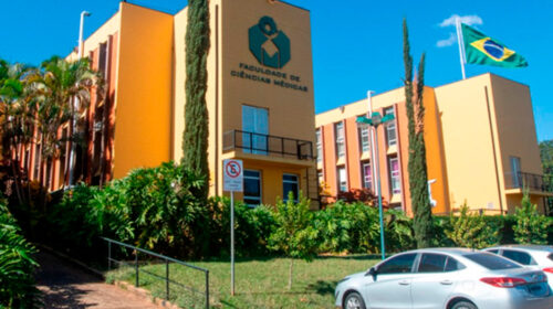 Fachada da Faculdade de Ciências Médicas (FCM) da Unicamp, com o prédio principal em destaque, vegetação ao redor e a bandeira do Brasil hasteada ao fundo.