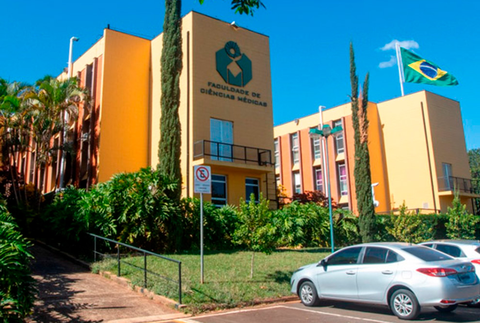 Fachada da Faculdade de Ciências Médicas (FCM) da Unicamp, com o prédio principal em destaque, vegetação ao redor e a bandeira do Brasil hasteada ao fundo.