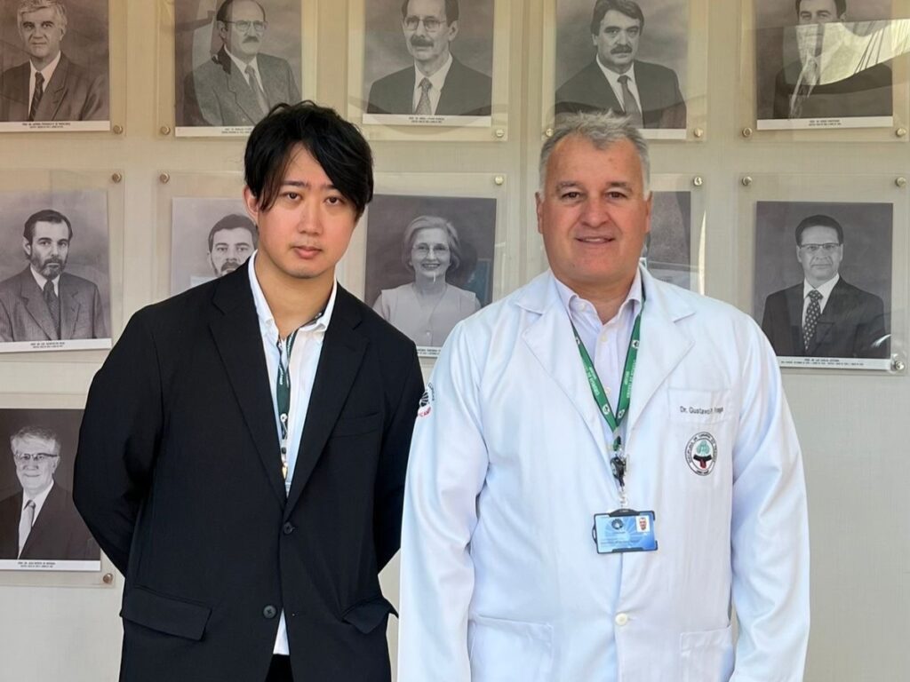 Dois homens posam para foto em frente a uma parede com retratos emoldurados em preto e branco. À esquerda, um jovem de terno preto e camisa branca; à direita, um homem de jaleco branco com crachá e cordão verde, identificado como médico.