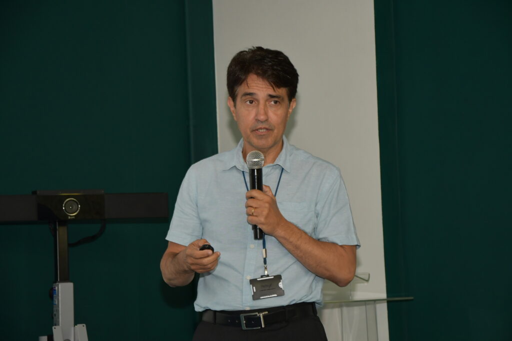 Homem em pé, segurando um microfone e um controle de apresentação enquanto fala em frente a uma parede verde e branca. Ele usa camisa social clara, crachá e cinto escuro.
