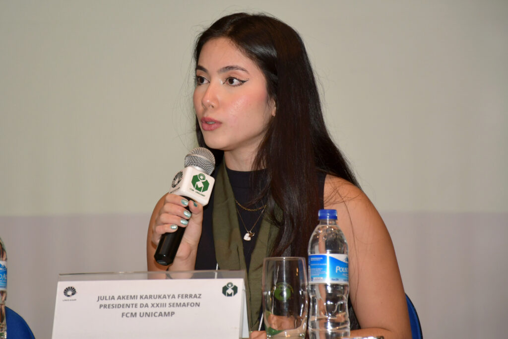 A estudante Julia Akemi Karukaya Ferraz fala ao microfone durante a abertura da 23ª Semana Acadêmica de Fonoaudiologia (Semafon) da FCM Unicamp. À sua frente, uma placa de identificação traz seu nome e o cargo de presidente da comissão organizadora.