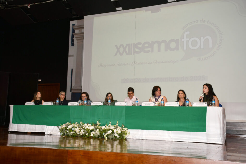 Mesa de abertura da 23ª Semana Acadêmica de Fonoaudiologia da FCM Unicamp, composta por docentes e convidados, posicionada sobre palco com toalha verde e arranjo de flores brancas. Ao fundo, o telão exibe o logotipo do evento e o período de realização, de 22 a 26 de setembro de 2025.