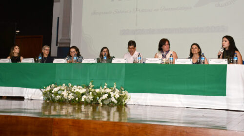 Mesa de abertura da 23ª Semana Acadêmica de Fonoaudiologia da FCM Unicamp, composta por docentes e convidados, posicionada sobre palco com toalha verde e arranjo de flores brancas. Ao fundo, o telão exibe o logotipo do evento e o período de realização, de 22 a 26 de setembro de 2025.