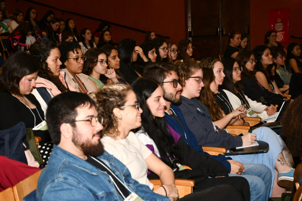 Público atento acompanha a programação da 23ª Semana Acadêmica de Fonoaudiologia da FCM Unicamp em auditório. A plateia é composta majoritariamente por estudantes, alguns fazendo anotações e outros ouvindo as apresentações.