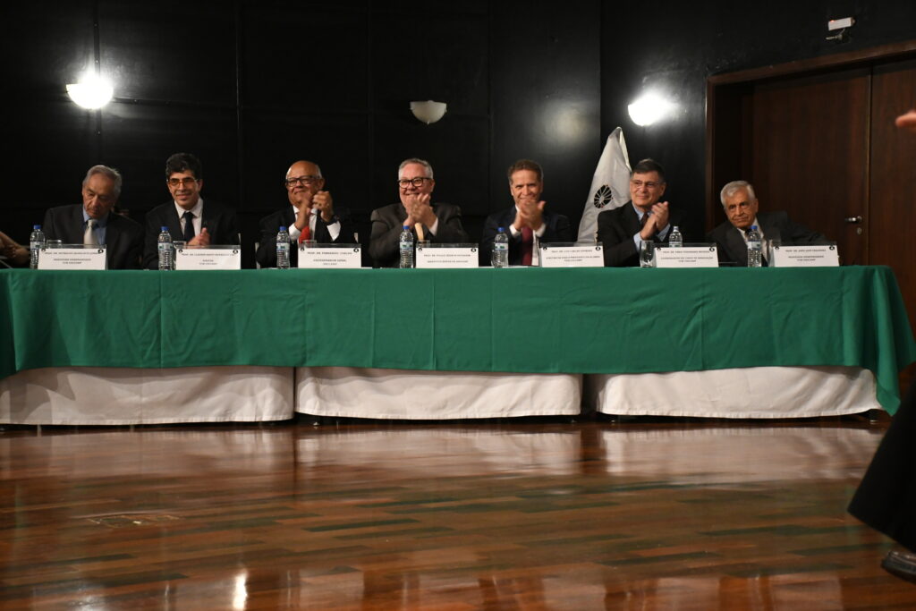 Integrantes da mesa diretiva da cerimônia dos 50 anos de formatura da 8ª Turma de Medicina da Unicamp estão sentados atrás de uma mesa coberta com toalha verde, aplaudindo no auditório; todos vestem trajes formais e há placas de identificação com seus nomes à frente.