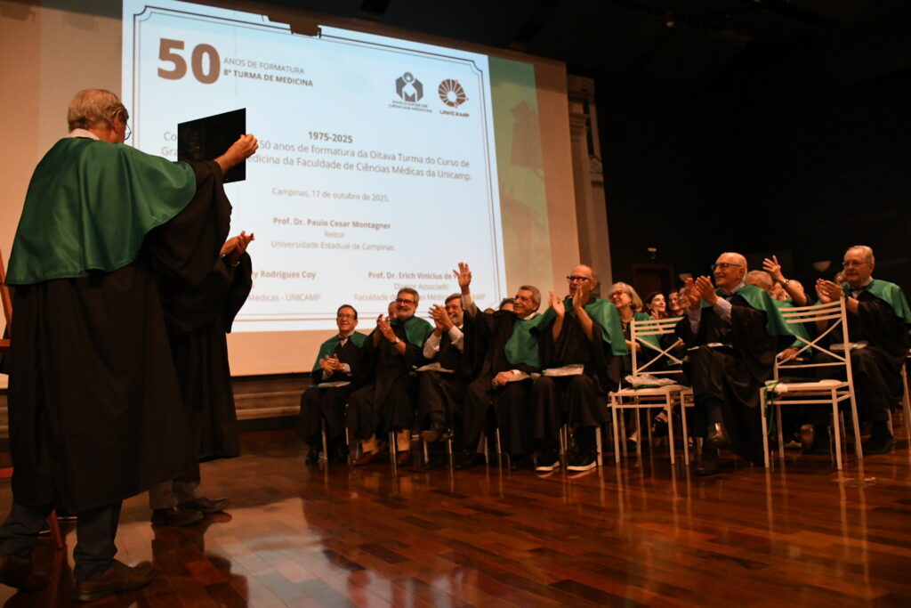 Formandos da 8ª Turma de Medicina aplaudem no palco do auditório enquanto um colega exibe uma placa comemorativa diante de um telão com a identidade visual do evento de 50 anos de formatura.