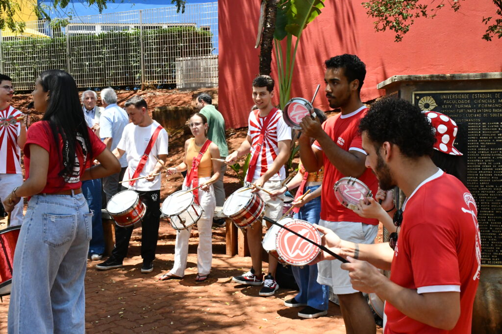 Grupo de estudantes da bateria universitária, com instrumentos de percussão, toca animadamente ao ar livre, próximo ao muro vermelho da Faculdade de Ciências Médicas da Unicamp, durante a recepção aos formandos.
