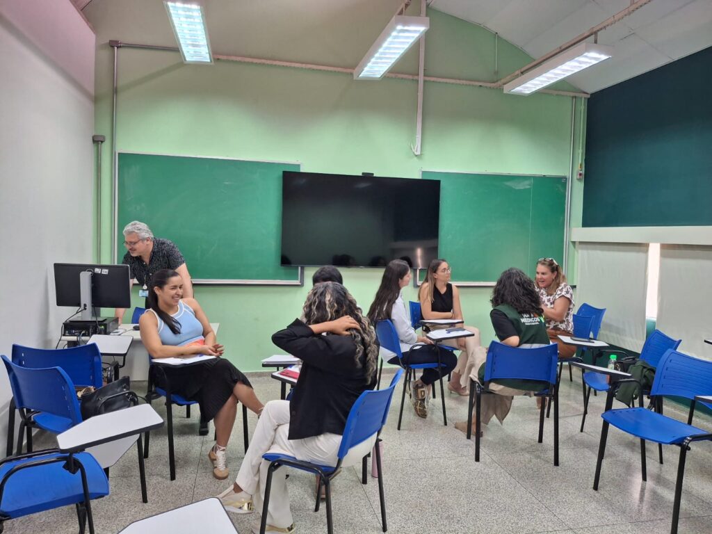 Grupo de pessoas em sala de aula, sentadas em círculo em cadeiras azuis, participando de roda de conversa.