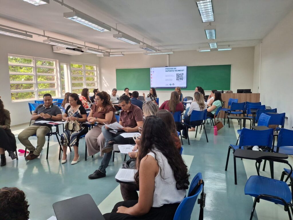 Sala com cadeiras azuis organizadas em círculo, ocupadas por pessoas em discussão, segurando folhas e materiais de apoio.