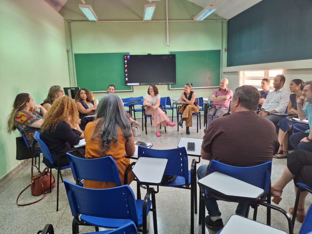 Pessoas em sala de aula, dispostas em roda de cadeiras azuis, interagindo em atividade de grupo.