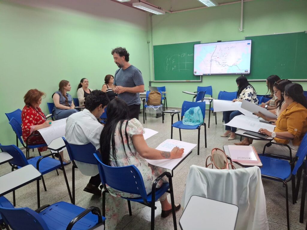 Participantes em sala de aula verde, sentados em círculo, atentos à discussão coletiva.