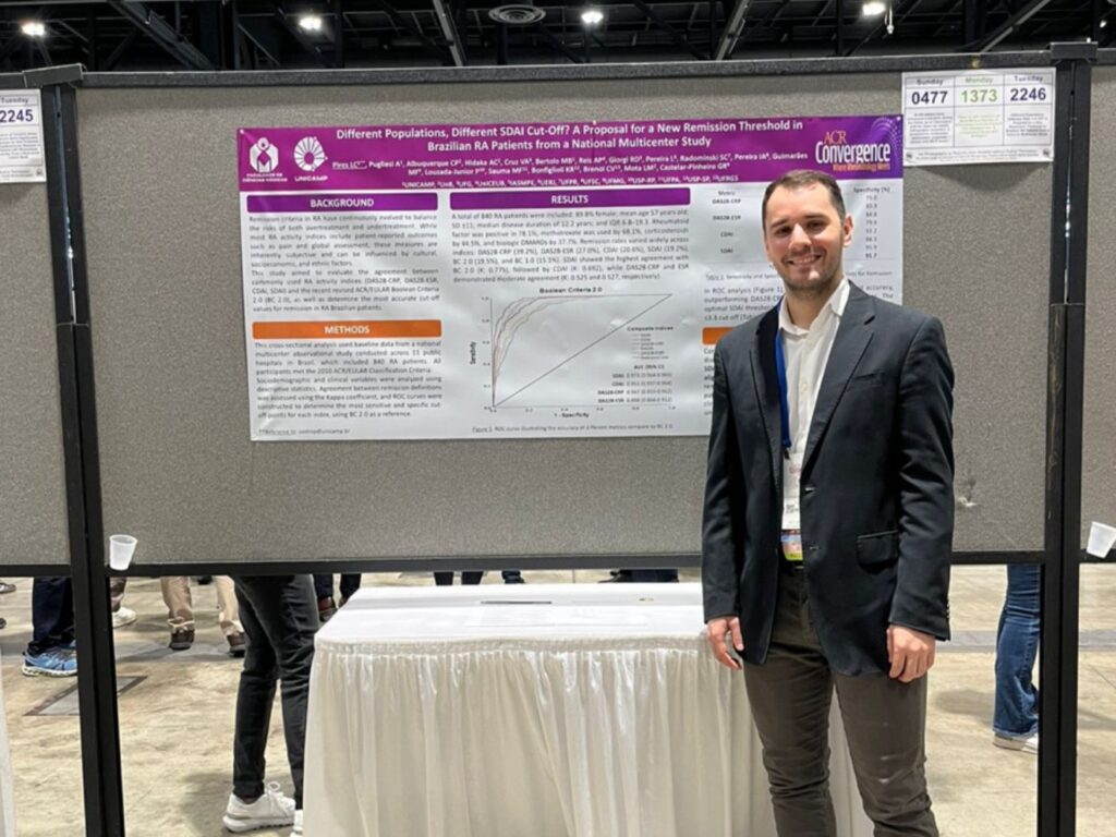 Homem de terno preto e camisa clara sorri ao lado de um pôster científico exposto em painel cinza durante o congresso ACR Convergence 2025. O pôster apresenta o título Different Populations, Different SDAI Cut-Off? A Proposal for a New Remission Threshold in Brazilian RA Patients from a National Multicenter Study, com logotipos da Unicamp e do congresso no topo.