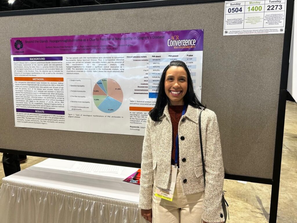 Mulher sorri em frente a um pôster científico fixado em painel cinza durante o congresso ACR Convergence 2025. O pôster, com logotipos da Unicamp e do congresso, tem o título Beyond the Glands: Hypogammaglobulinemia as a Clue to Peripheral Neuropathy in Sjogren’s Disease e exibe um gráfico de setores colorido no centro.