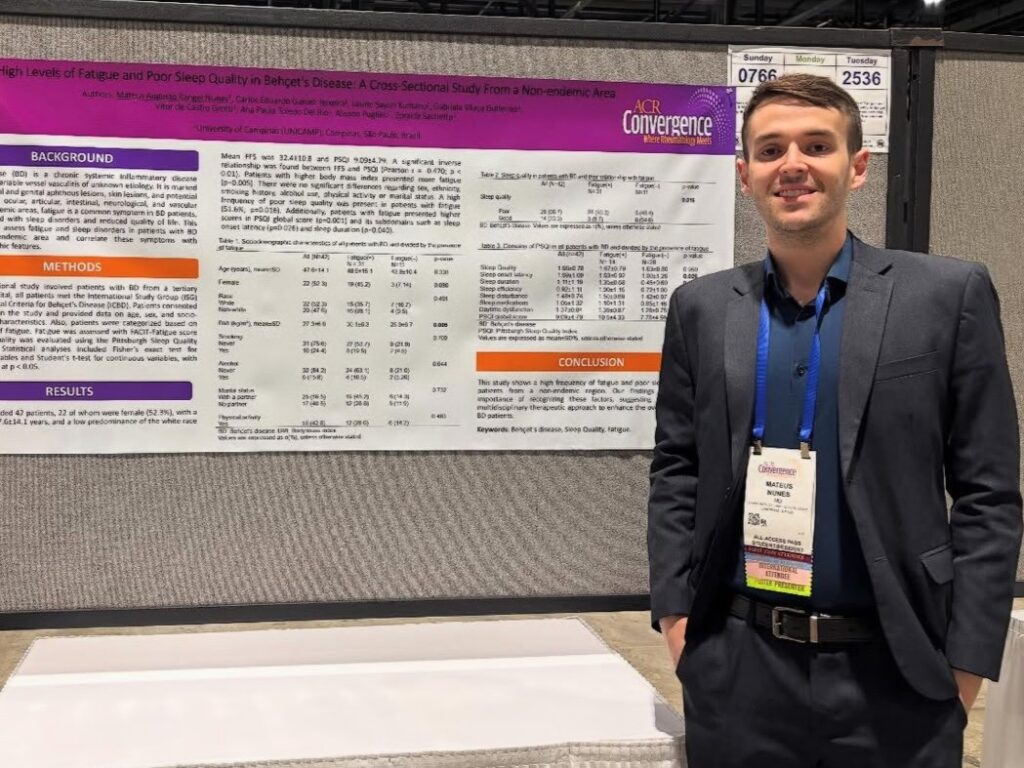 Homem veste blazer escuro e camisa azul e posa sorridente diante de um pôster científico apresentado no congresso ACR Convergence 2025. O pôster tem o título High Levels of Fatigue and Poor Sleep Quality in Behçet’s Disease: A Cross-Sectional Study from a Non-endemic Area, com logotipos da Unicamp e do congresso no topo.