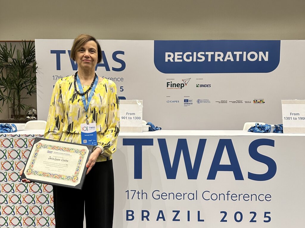 A terceira foto apresenta a Professora Iscia Lopes Cendes posando em frente à área de REGISTRATION (Inscrição) da 17ª Conferência Geral da TWAS, Brasil 2025. Ela está sorridente e segura um certificado ou diploma emoldurado, com a legenda da foto indicando que ela foi eleita em 2024 e empossada no congresso de 2025 da TWAS. A professora usa uma blusa amarela e preta estampada, um crachá da conferência e calças pretas. O fundo mostra a marca e os patrocinadores (Finet, BNDES, CAPES, UNESCO) do evento.