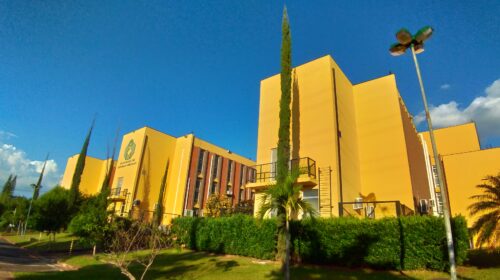 Prédios amarelos da Faculdade de Ciências Médicas da Unicamp vistos de ângulo lateral, com o logotipo da unidade à esquerda, vegetação e árvores altas em frente, postes de iluminação e céu azul com algumas nuvens ao fundo.