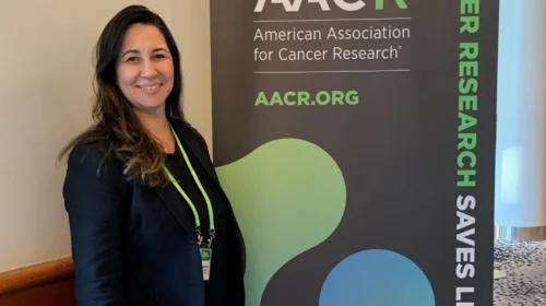 Mulher em pé ao lado de um banner da conferência da American Association for Cancer Research (AACR). Ela sorri para a câmera e usa blazer preto e calça azul. O banner exibe o logotipo da AACR e o texto “Cancer Research Saves Lives”.