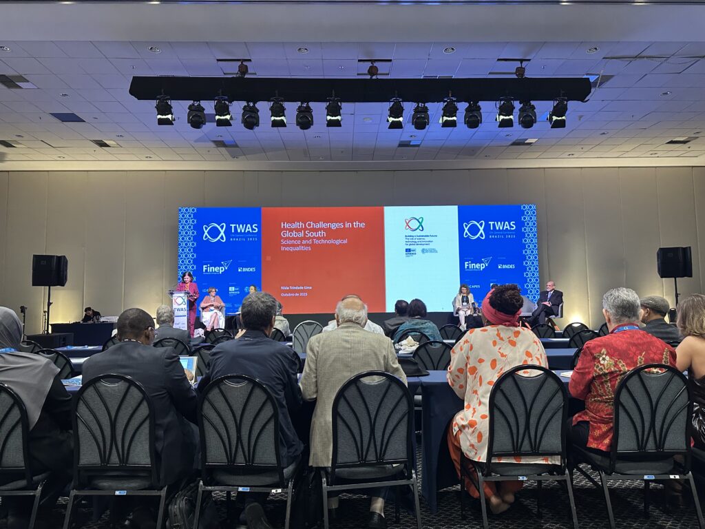 A segunda imagem captura uma visão mais ampla do Simpósio sobre Saúde no Sul Global, parte da 17ª Conferência Geral da TWAS no Brasil. O palco está iluminado, com painéis de LED exibindo o título do simpósio, "Health Challenges in the Global South" (Desafios de Saúde no Sul Global), e a menção a "Science, Technology, and Inequalities" (Ciência, Tecnologia e Desigualdades). Um painelista está sentado à direita, enquanto duas pessoas — uma delas provavelmente a Professora Helena Nader — estão nos púlpitos à esquerda. A plateia é vista de costas, sentada em fileiras, assistindo à discussão. A iluminação de palco e as grandes caixas de som laterais indicam que o evento é realizado em um grande centro de conferências.