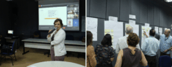 Em uma sala de reunião, uma mulher segura um microfone e fala diante de uma projeção que exibe tópicos e participantes em videoconferência, enquanto, ao lado, um grupo de pessoas interage com murais preenchidos com cartazes e post-its organizados por categorias como “Perspectiva Cliente e Sociedade”, “Processos” e “Gestão”.