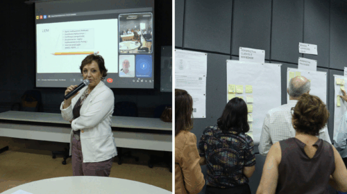 Em uma sala de reunião, uma mulher segura um microfone e fala diante de uma projeção que exibe tópicos e participantes em videoconferência, enquanto, ao lado, um grupo de pessoas interage com murais preenchidos com cartazes e post-its organizados por categorias como “Perspectiva Cliente e Sociedade”, “Processos” e “Gestão”.