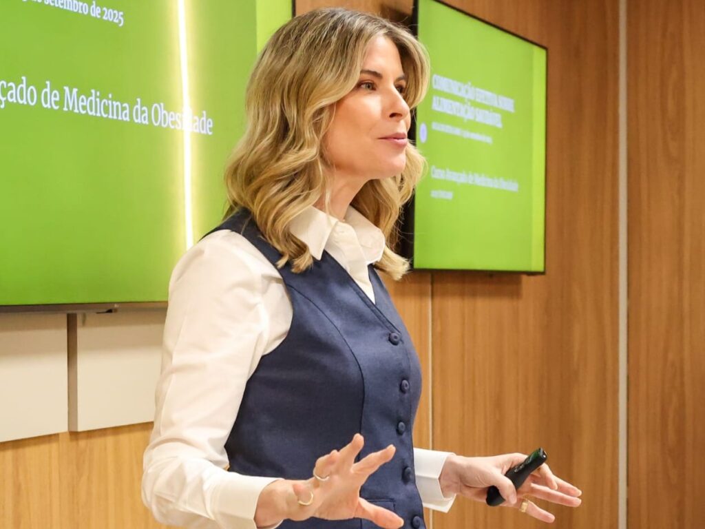 Close-up de Rita Lobo gesticulando com as mãos durante sua apresentação. Ela está em frente a telas de projeção que mencionam "Comunicação Efetiva" e temas relacionados à "Medicina da Obesidade".