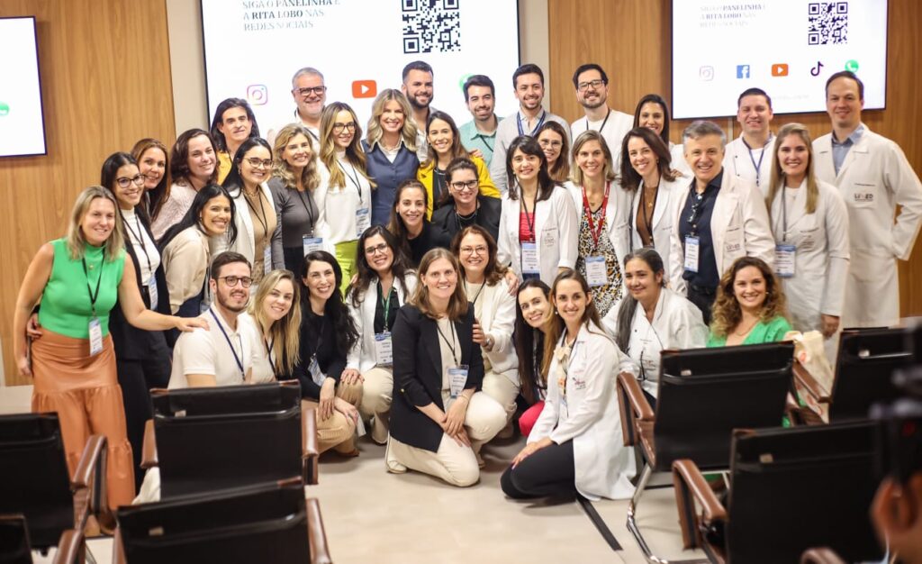 Foto de grupo grande no auditório. Dezenas de pessoas de jaleco posam com os palestrantes. Uma tela ao fundo exibe referências a Panelinha e Rita Lobo nas redes sociais.
