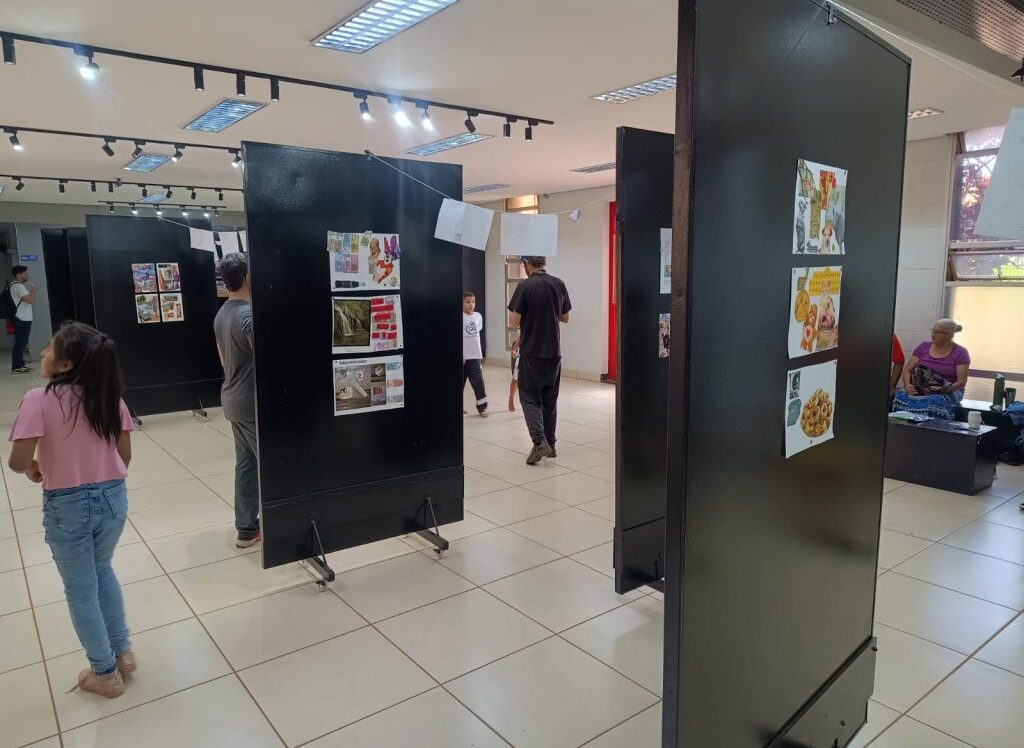 Visitantes observam painéis de uma exposição montada em um espaço interno amplo e bem iluminado, com piso claro e teto com luminárias direcionadas. As paredes exibem fotos e textos, e há adultos e crianças circulando pelo local. Ao fundo, uma porta de vidro com a placa “Espaço Cultural” indica a entrada do ambiente.