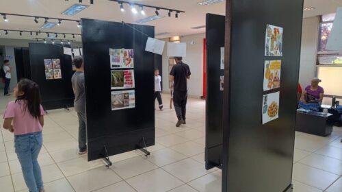 Visitantes observam painéis de uma exposição montada em um espaço interno amplo e bem iluminado, com piso claro e teto com luminárias direcionadas. As paredes exibem fotos e textos, e há adultos e crianças circulando pelo local. Ao fundo, uma porta de vidro com a placa “Espaço Cultural” indica a entrada do ambiente.