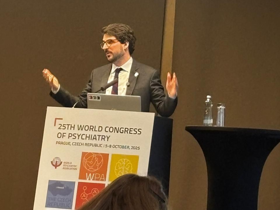 Um homem de terno e gravata faz uma apresentação no púlpito durante o 25th World Congress of Psychiatry, realizado em Praga, República Tcheca. Ele gesticula com as mãos enquanto fala, e à frente do púlpito há um painel com o logotipo e informações do congresso.