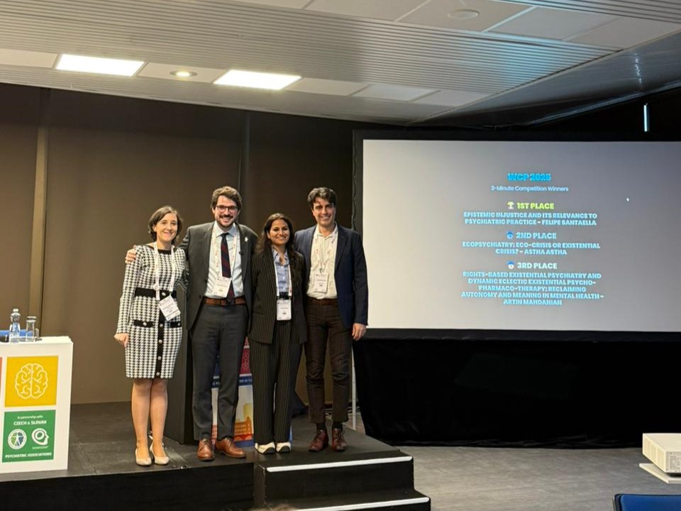 Quatro pessoas posam sorridentes no palco após a premiação do 25th World Congress of Psychiatry. Ao fundo, uma tela projeta o resultado da competição “3-Minute Competition Winners”, destacando o primeiro lugar conquistado por Felipe Santaella.