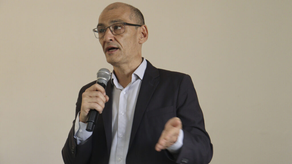 Close-up de um homem calvo de óculos, vestindo terno escuro e camisa clara, falando ao microfone. Sua mão direita está erguida, gesticulando.