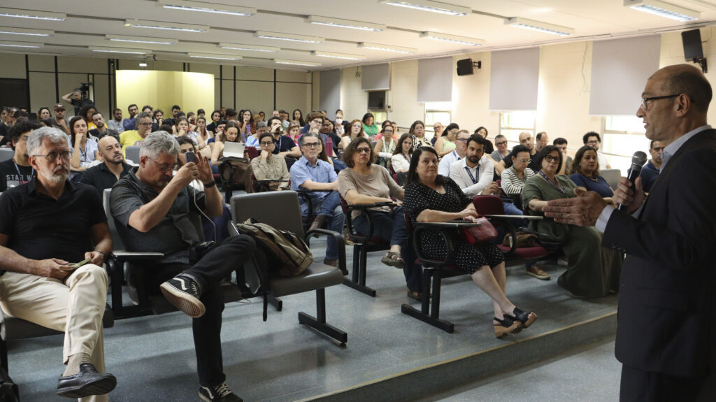 Visão ampla de um auditório com uma grande plateia sentada. O mesmo homem de terno do arquivo a14 está em pé à direita, falando ao microfone para os participantes.