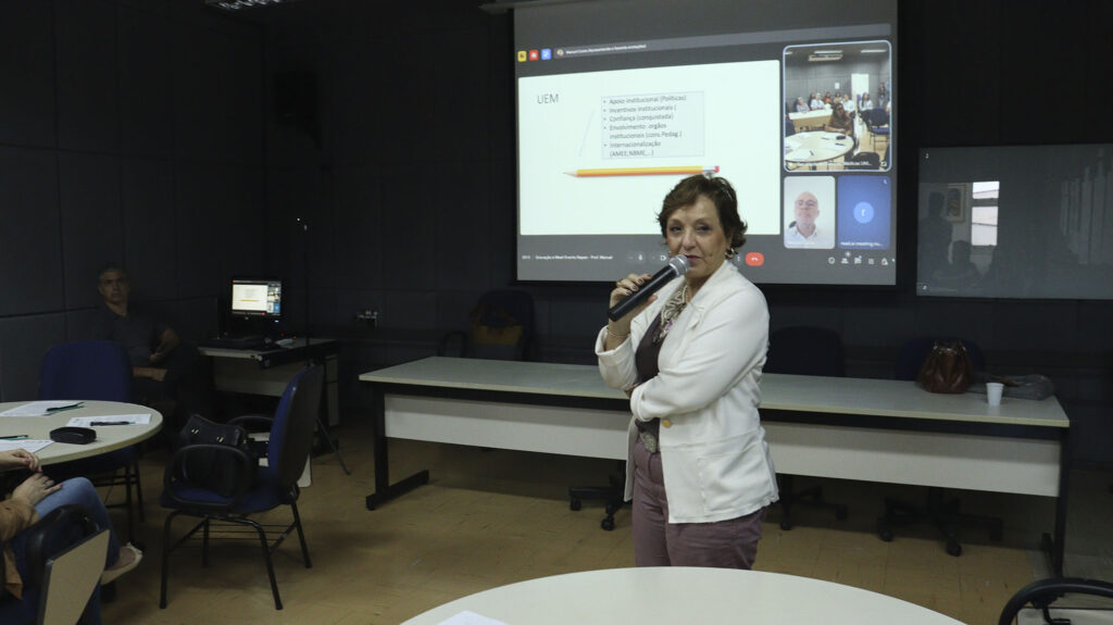 Mulher em pé, segurando um microfone, fala para os participantes em uma sala de reuniões. Ao fundo, projeção de uma apresentação e chamada de videoconferência na tela.