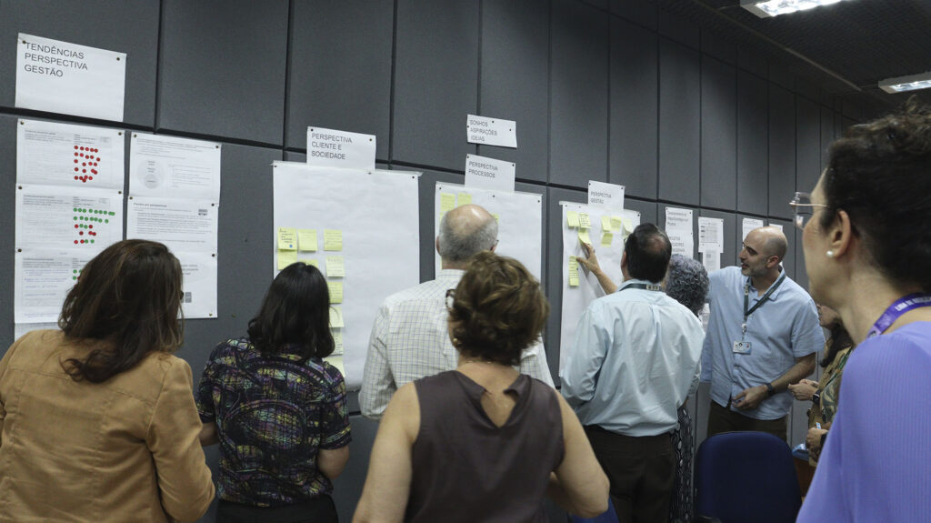 Grupo de pessoas em pé diante de painéis na parede, interagindo com cartazes e anotações em post-its organizados por temas como “Gestão”, “Processos” e “Sociedade”.