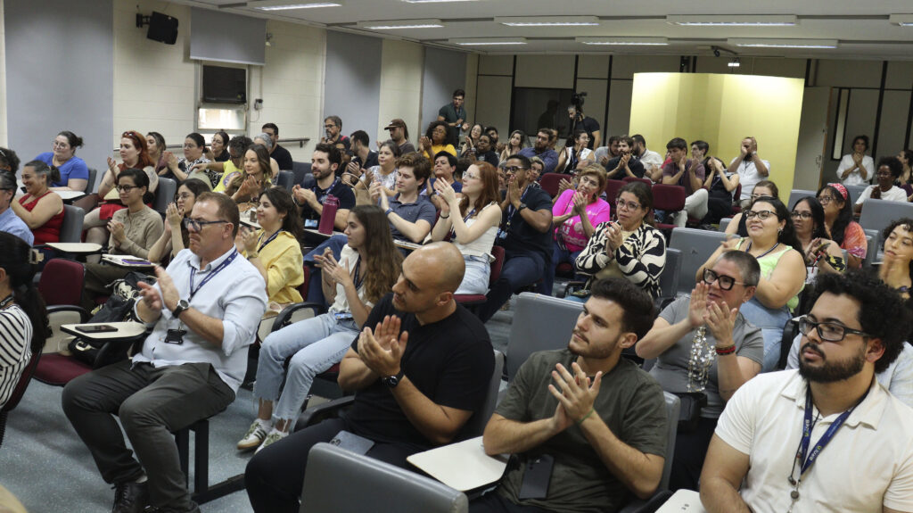 Plateia de um auditório com dezenas de pessoas visivelmente engajadas e aplaudindo a apresentação.