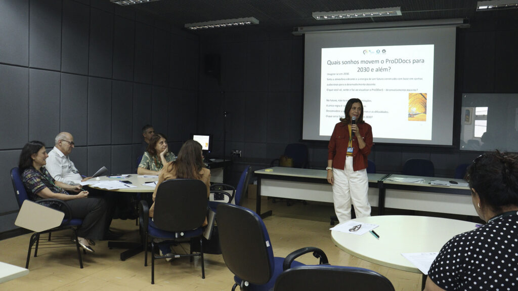 Mulher em pé, segurando um microfone, fala para um grupo de pessoas sentadas em mesas redondas. Ao fundo, apresentação projetada com a pergunta “Quais sonhos movem o ProDDocs para 2030 e além?”.