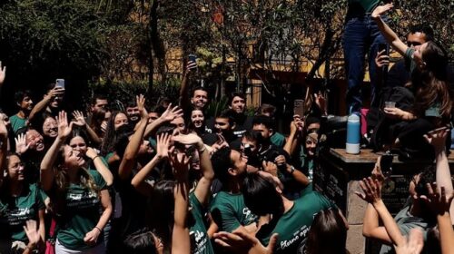 Um grande grupo de jovens, muitos vestindo camisetas verdes, comemora ao ar livre sob o sol. Uma pessoa em destaque está em pé sobre uma base de pedra, segurando o celular para tirar uma selfie enquanto sorri, cercada por colegas que levantam os braços e também registram o momento. Ao fundo, há árvores e construções amarelas. A cena transmite alegria e celebração coletiva.