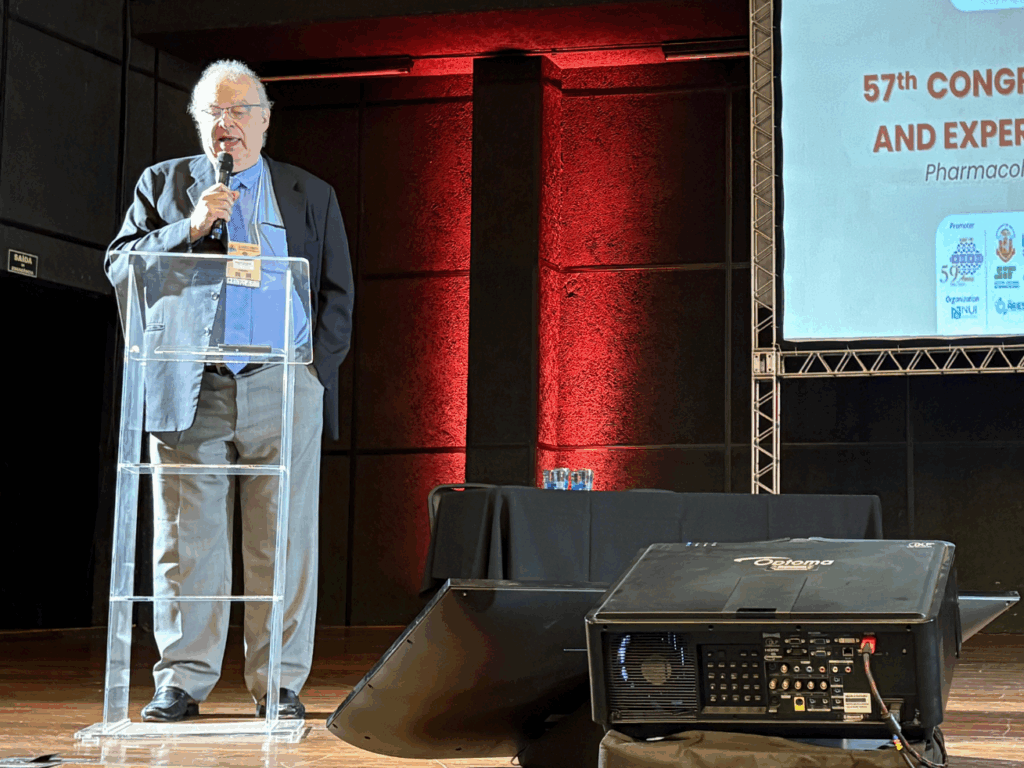 O professor Gilberto de Nucci fala ao microfone em um púlpito transparente durante o 57º Congresso Brasileiro de Farmacologia e Terapêutica Experimental, com projeção de slide ao fundo e iluminação avermelhada no palco.