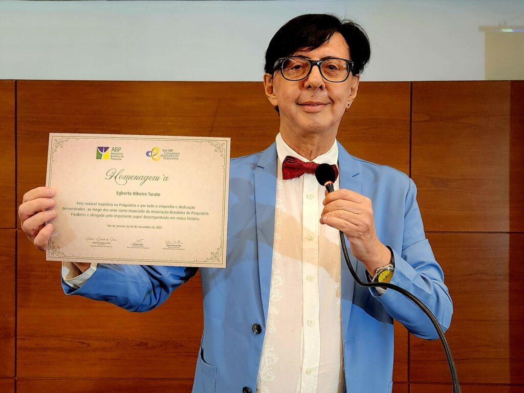 Homem de terno azul-claro, camisa branca e gravata borboleta vermelha segura certificado de homenagem da Associação Brasileira de Psiquiatria. Ele está em pé diante de um painel de madeira, sorrindo levemente.