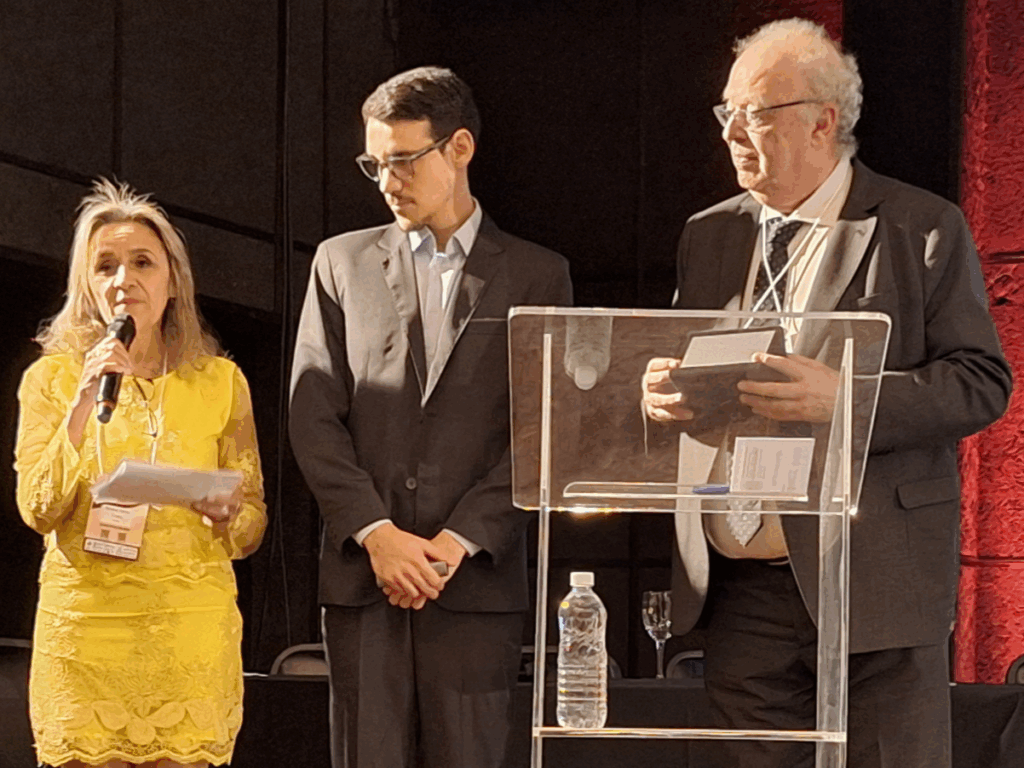 A presidente da SBFTE, Soraia Costa, fala ao microfone ao lado de um jovem e do professor Gilberto de Nucci, que segura uma placa de premiação no palco do congresso.