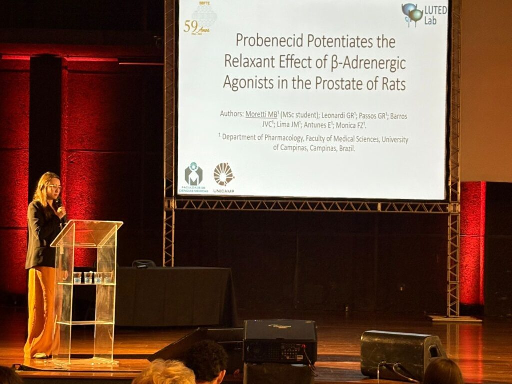 A estudante Mariana faz apresentação de seu trabalho no palco, em frente a um púlpito transparente, com o slide projetado exibindo o título do estudo e os logotipos da Faculdade de Ciências Médicas e da Unicamp.