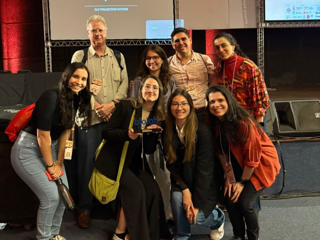 Parte da equipe da Faculdade de Ciências Médicas da Unicamp posa para foto em grupo no auditório do congresso, com oito pessoas sorrindo diante do palco e de equipamentos de projeção.